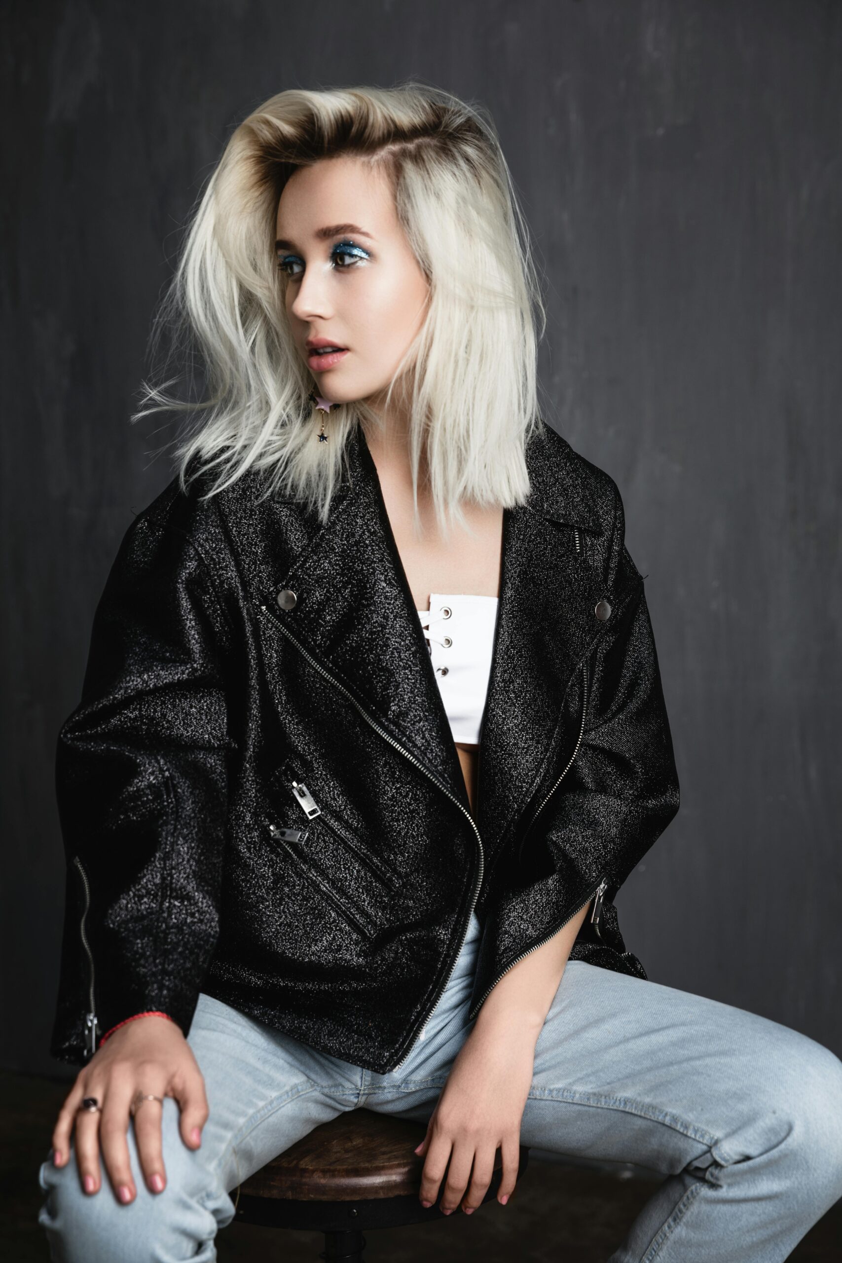 Girl with blonde hair sitting sideways on na stool with a black leather biker jacket on