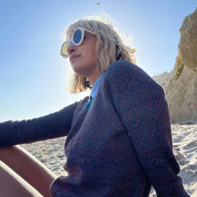 Blonde beach hair girl with white sunglasses and a nose piercing sitting on a beach, with a glow of sunshine right behind her.